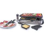 Raclette shown with food on top of grill, melted cheese in raclette dishes in front of raclette and a plate full of food to the left